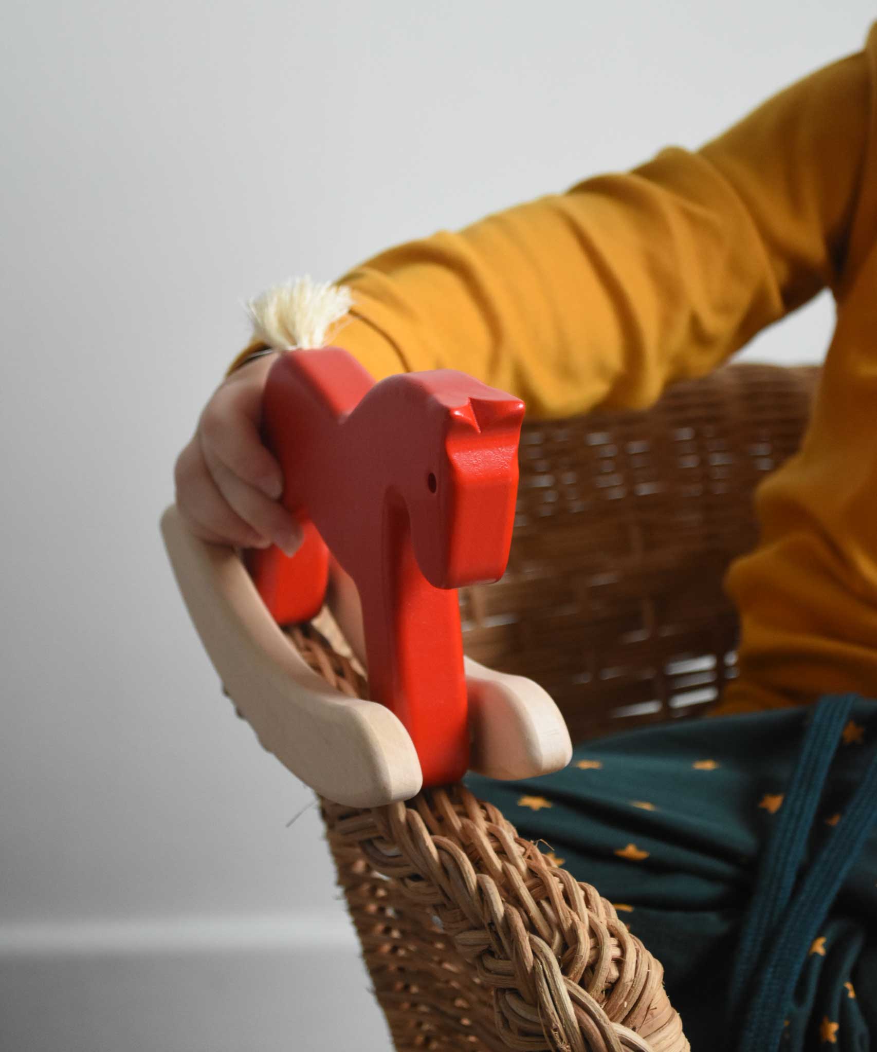 A close up of a child's hand holding a red coloured Bajo Mini Rocking Horse. The child is sitting in a wicker chair. These heirloom quality toys by Bajo are part of a wide range of wooden toys available here at Babipur. 