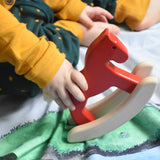 A close up of a child's hand holding a red coloured Bajo Mini Rocking Horse. The child is sitting on a Wonderie farm themed wonder cloth. These heirloom quality toys by Bajo are part of a wide range of wooden toys available here at Babipur. 