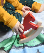 A close up of a child's hand holding a red coloured Bajo Mini Rocking Horse. The child is sitting on a Wonderie farm themed wonder cloth. These heirloom quality toys by Bajo are part of a wide range of wooden toys available here at Babipur. 
