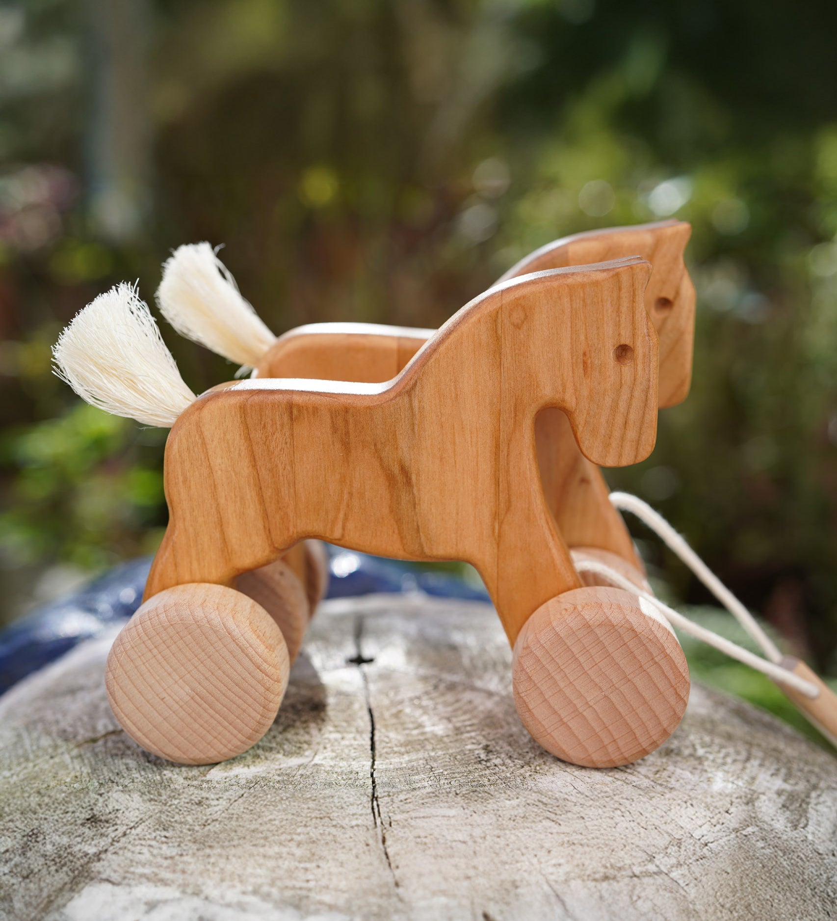 Bajo hand made wooden natural jumping horse available at Babipur. Features unpainted wheels and a cut out hole for easy grabbing. Horses are standing on a wooden surface in a outdoor setting