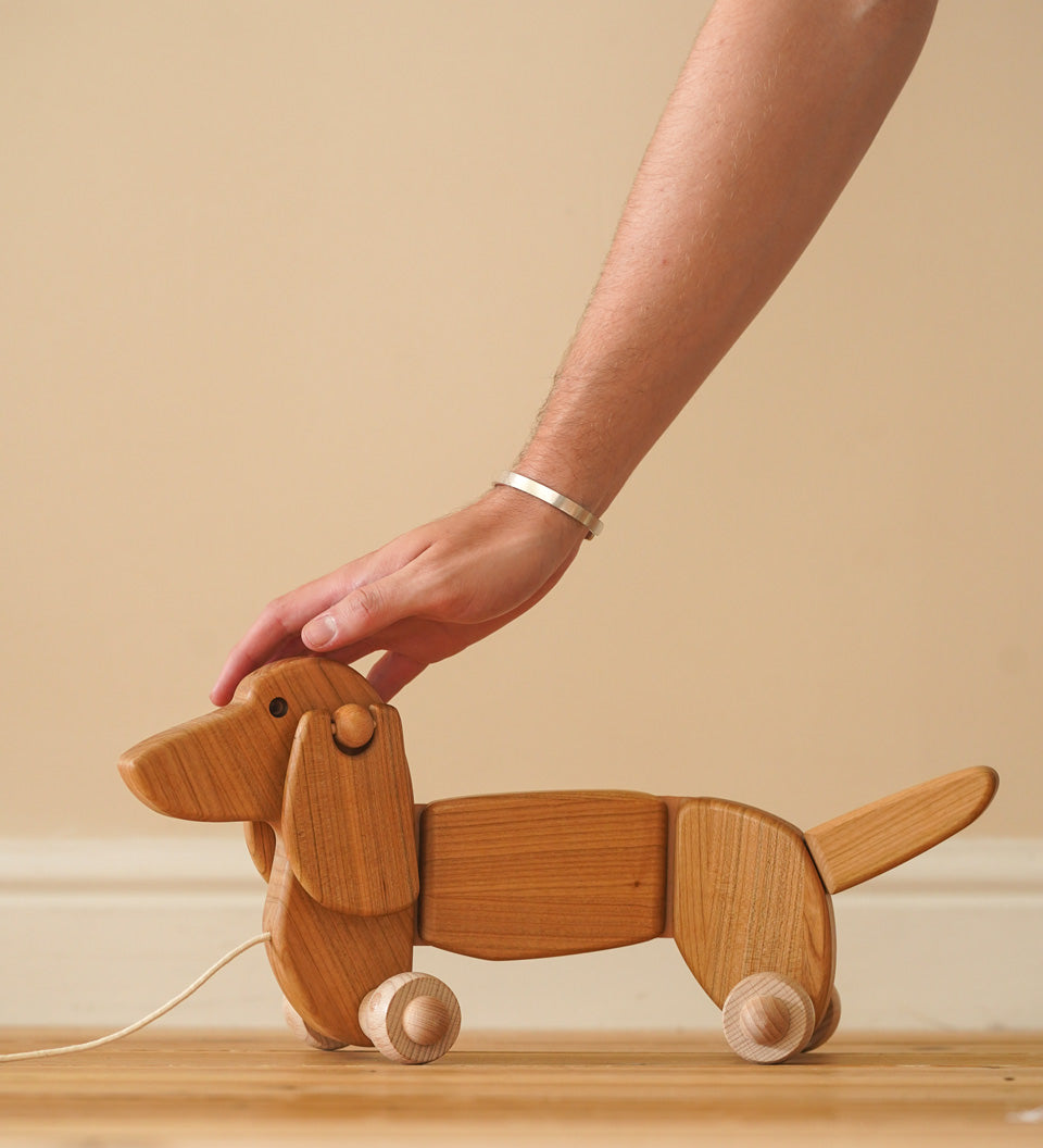 Close up of a hand reaching down to touch the Bajo natural wooden toy dachshund on a wooden floor