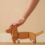 Close up of a hand reaching down to touch the Bajo natural wooden toy dachshund on a wooden floor