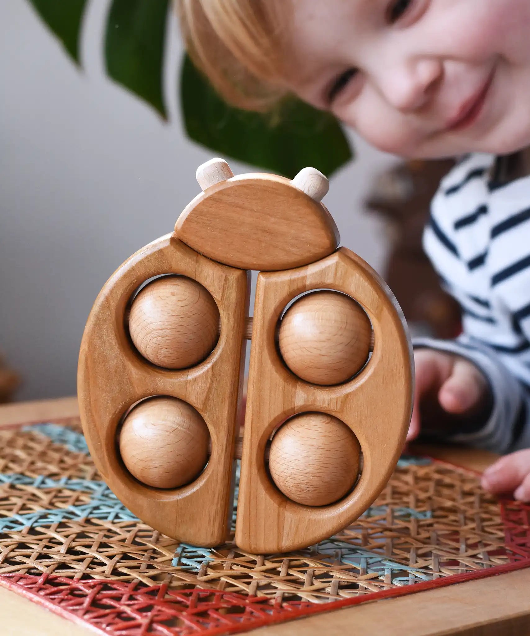 Bajo natural wooden ladybird teether  placed on a rattan stool in the Babipur playroom with a child looking on in the background.