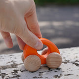 Adult hand pushing a Bajo handmade wooden orange ellipse car on a concrete surface showing scale