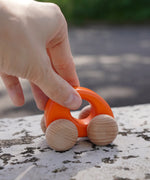 Adult hand pushing a Bajo handmade wooden orange ellipse car on a concrete surface showing scale