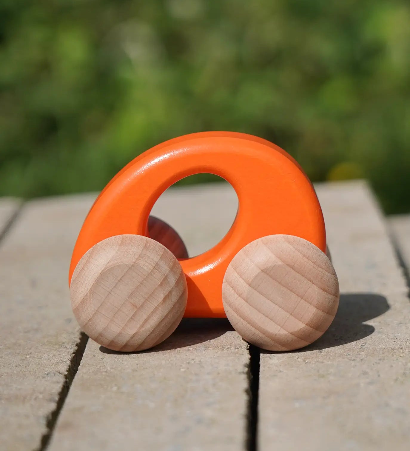 Bajo handmade wooden ellipse toy car in orange with unpainted wheels standing on a concrete surface