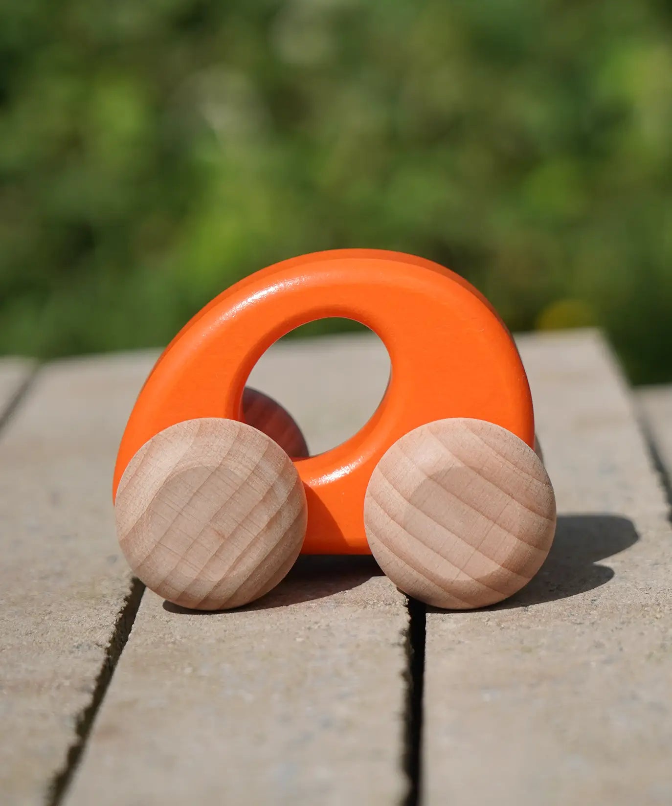Bajo handmade wooden ellipse toy car in orange with unpainted wheels standing on a concrete surface