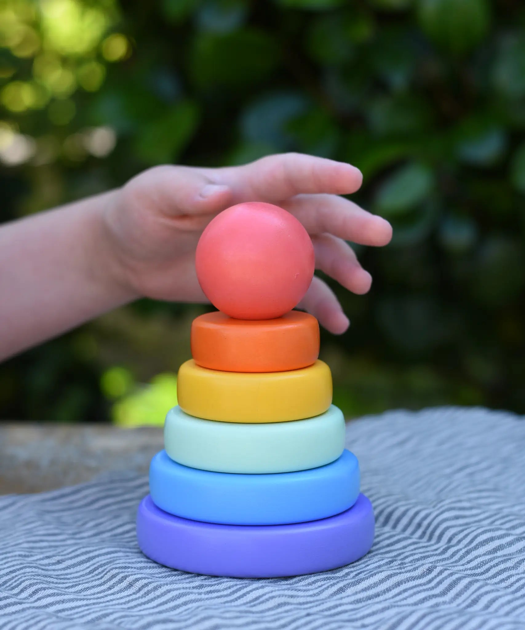 Child's hand reaching for the top ball shaped pieced from the Bajo pastel stacking rings wooden toy. 