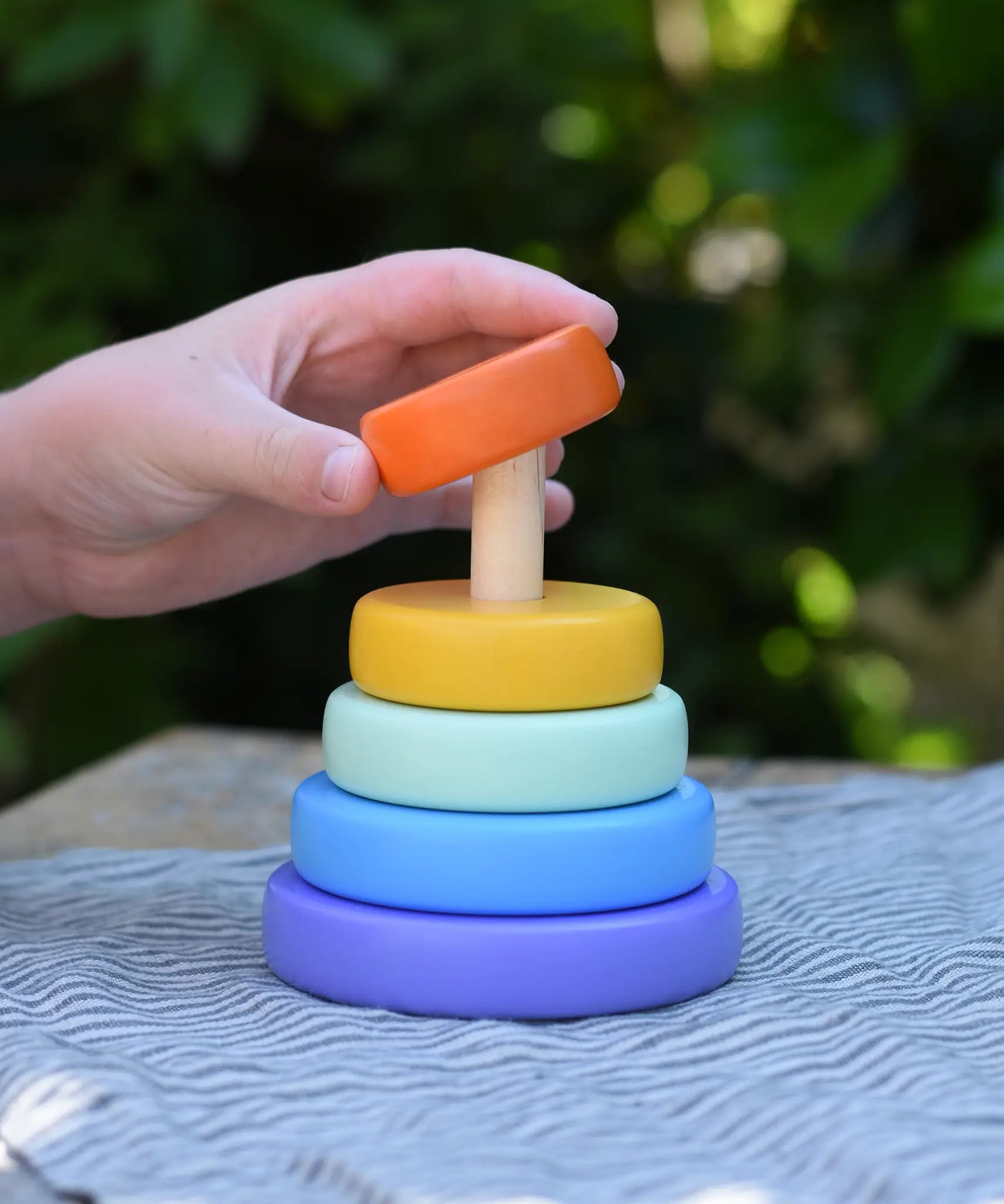 Child hand placing the orange ring onto the Bajo pastel stacking rings wooden toy. 