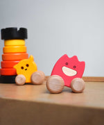A pink coloured Bajo wooden ghost toy placed on Triclimb Miri slide. A yellow ghost and a red rook tower stacking toy can be seen in the background.  The toy has a pink painted body and natural wooden wheels. These heirloom quality toys by Bajo are part of a wide range of wooden toys available here at Babipur. 