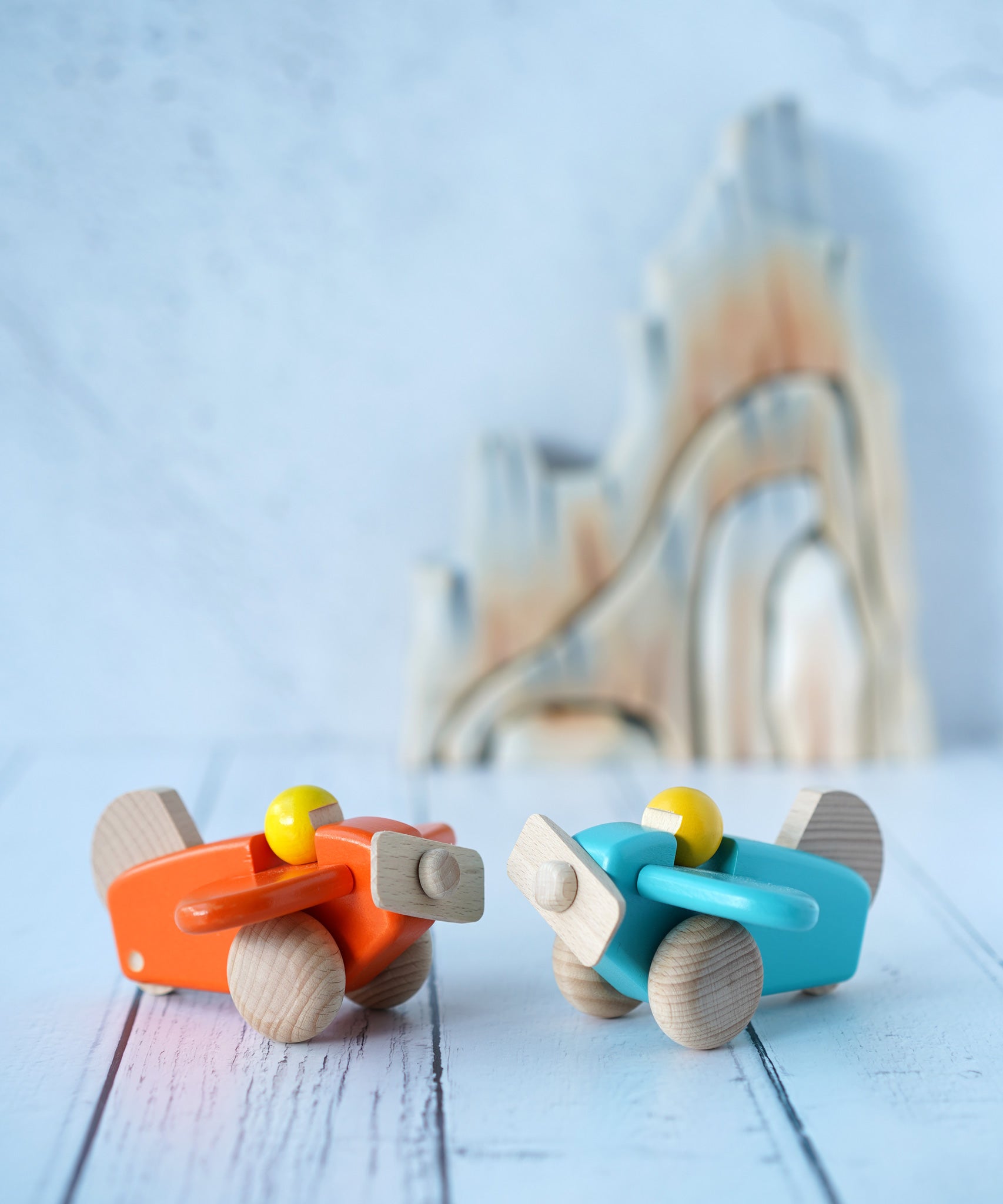 Two Bajo toy planes with pilots placed on a wooden surface in front of Bumbu rocks. One plane  This plane is part of a wide range of Bajo wooden toy vehicles available at Babipur.s orange the other is blue.