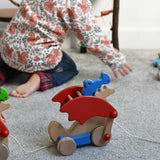 A Bajo blue pull along dragon wooden toy by the string across a Triclimb Miri slide.  The dragon toy features two wheels at the front and back and red wing that move along with the front wheels. A child can be seen playing in the background. These heirloom quality toys by Bajo are part of a wide range of wooden toys available here at Babipur.