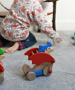 A Bajo blue pull along dragon wooden toy by the string across a Triclimb Miri slide.  The dragon toy features two wheels at the front and back and red wing that move along with the front wheels. A child can be seen playing in the background. These heirloom quality toys by Bajo are part of a wide range of wooden toys available here at Babipur.