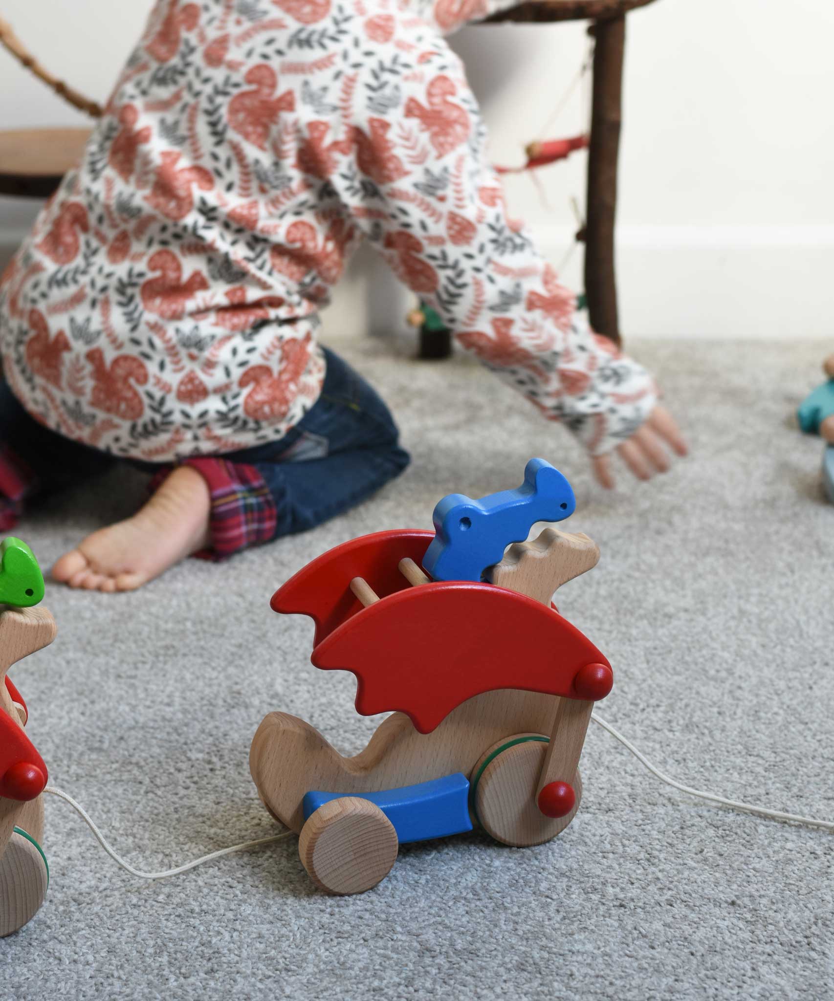 A Bajo blue pull along dragon wooden toy by the string across a Triclimb Miri slide.  The dragon toy features two wheels at the front and back and red wing that move along with the front wheels. A child can be seen playing in the background. These heirloom quality toys by Bajo are part of a wide range of wooden toys available here at Babipur.