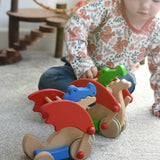 A child playing with the Bajo green pull along dragon wooden toy, the blue dragon has been placed in front of them. The dragon toy features two wheels at the front and back and red wing that move along with the front wheels. These heirloom quality toys by Bajo are part of a wide range of wooden toys available here at Babipur.