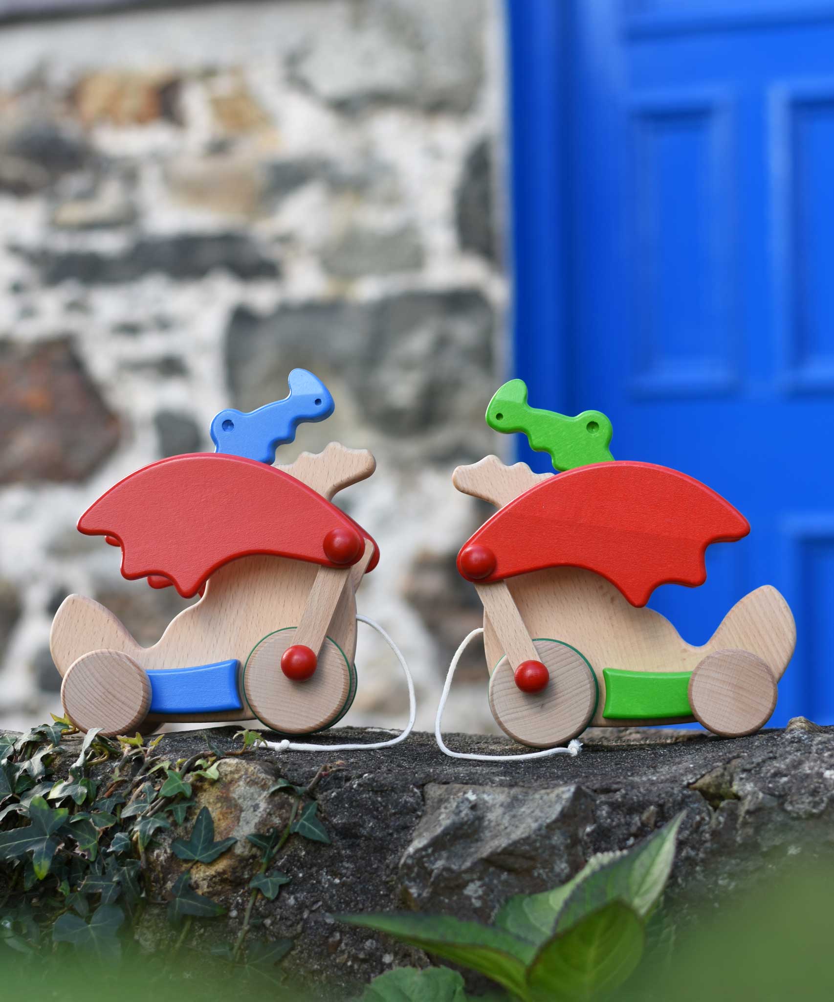 One blue and one green Bajo pull along dragon toys placed on a stone wall outdoors. These heirloom quality toys by Bajo are part of a wide range of wooden toys available here at Babipur. 