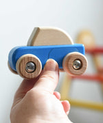 A blue coloured Bajo pull back action wooden toy car held up in an adult's hand. These heirloom quality toys by Bajo are part of a wide range of wooden toys available here at Babipur. 