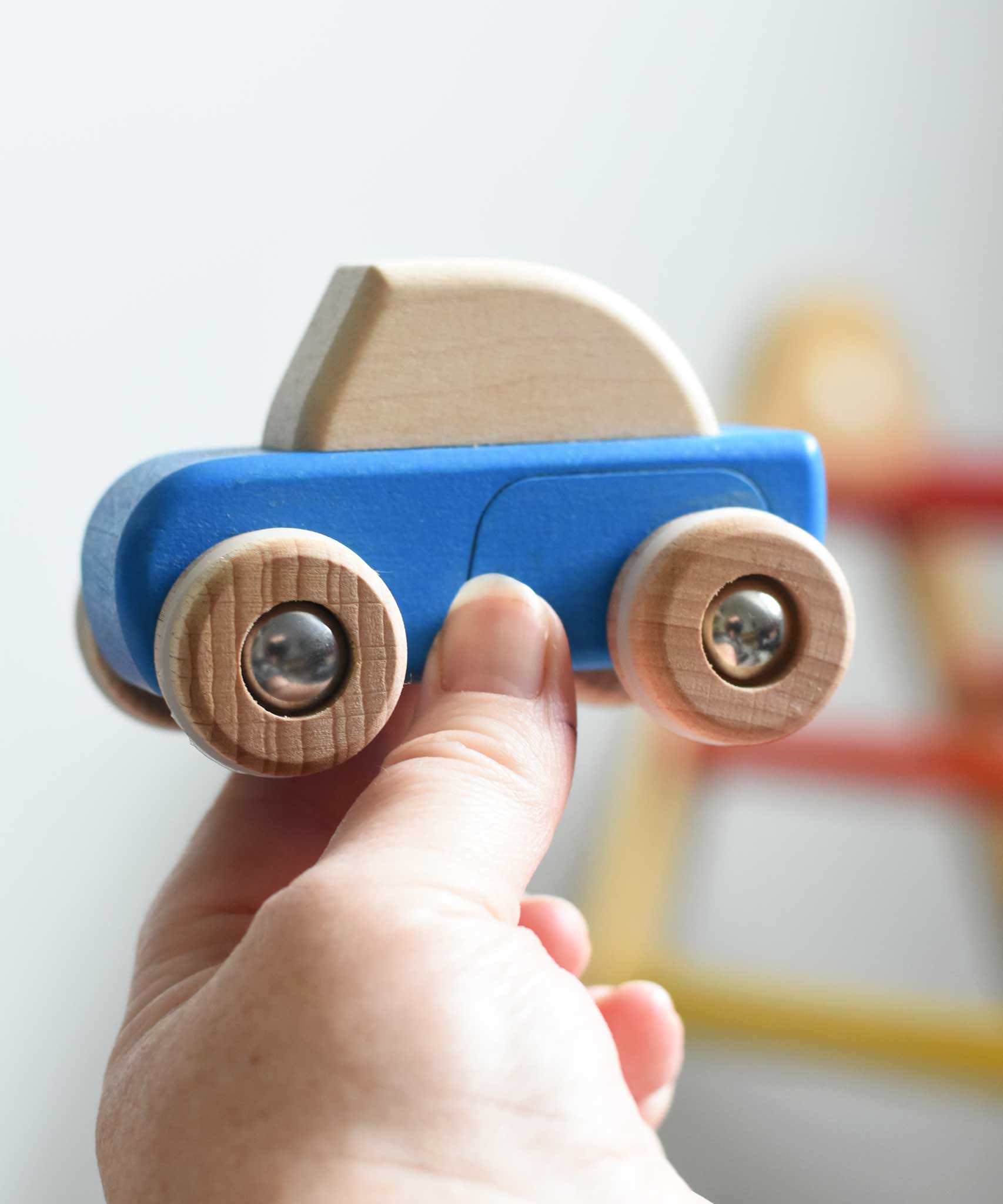 A blue coloured Bajo pull back action wooden toy car held up in an adult's hand. These heirloom quality toys by Bajo are part of a wide range of wooden toys available here at Babipur. 