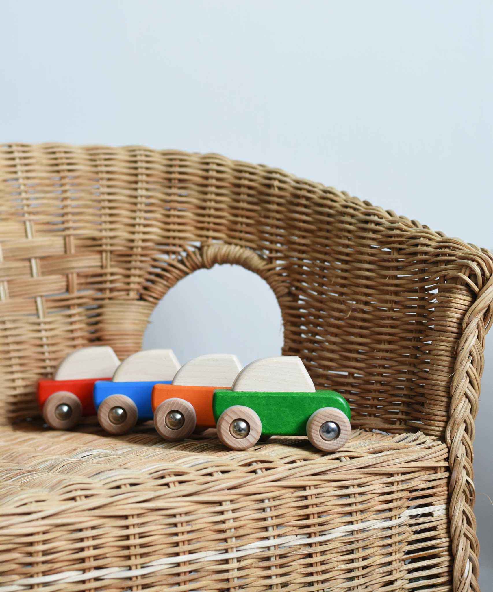 Four Bajo pull back action wooden toy cars placed on a child's wicker chair. These heirloom quality toys by Bajo are part of a wide range of wooden toys available here at Babipur. 