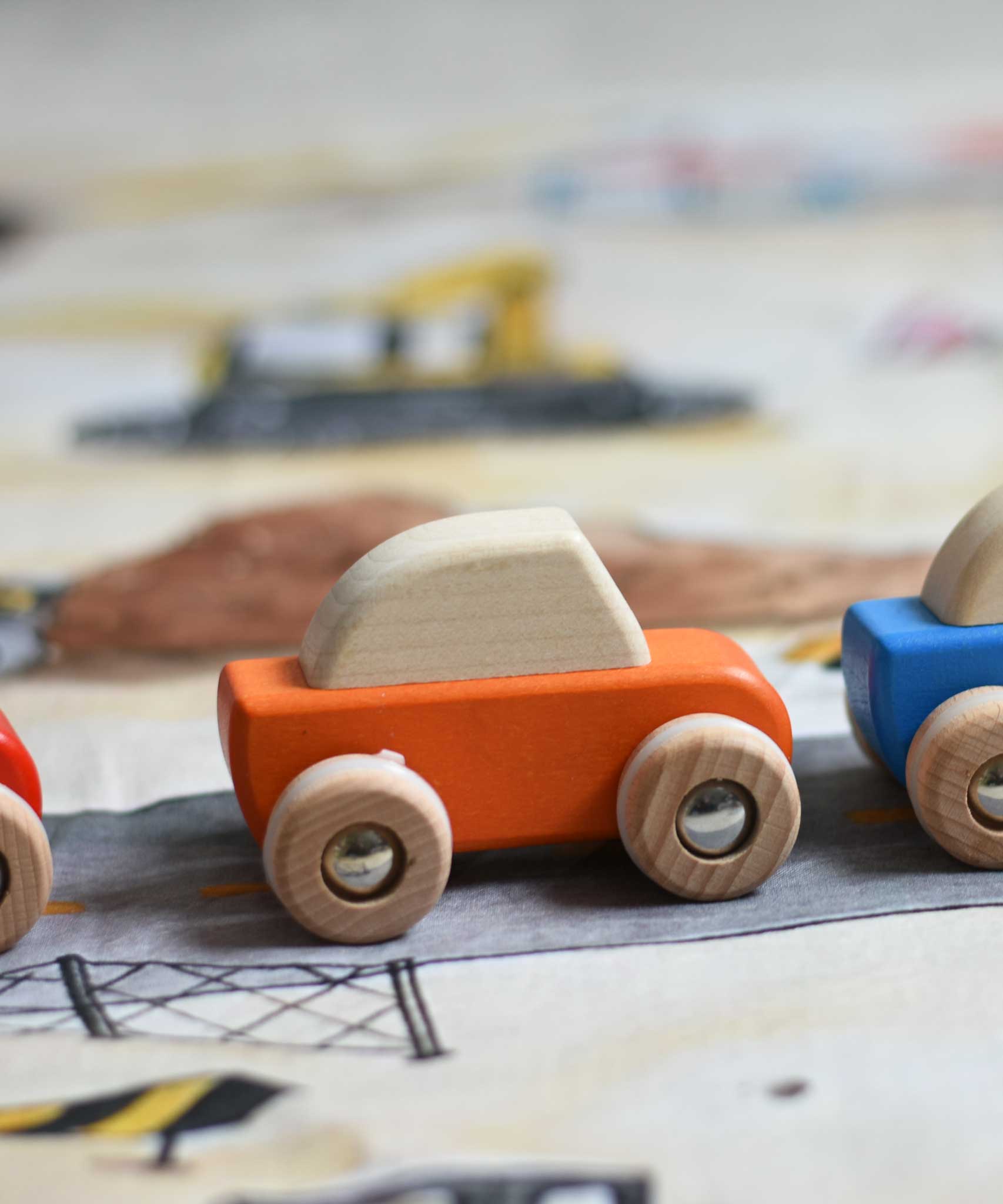 A close up of the orange coloured Bajo pull back action wooden toy car placed on the road printed onto the building site Wonderie wonder cloth. These heirloom quality toys by Bajo are part of a wide range of wooden toys available here at Babipur. 