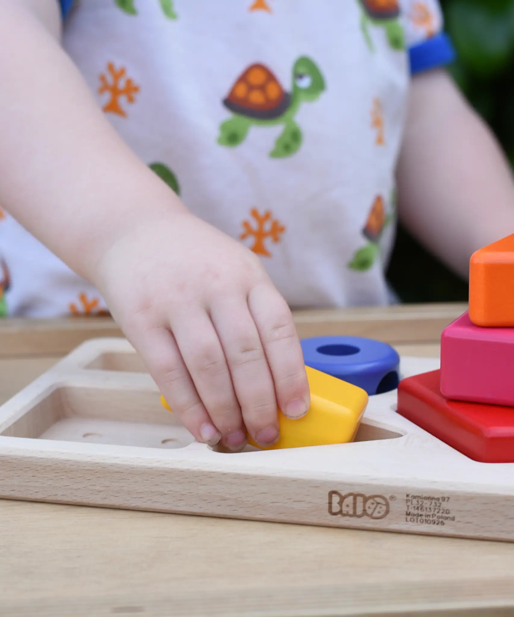 Child playing with the Bajo pyramid sorting board and stacker toy placing a yellow rectangle piece in the wooden base board frame. 
