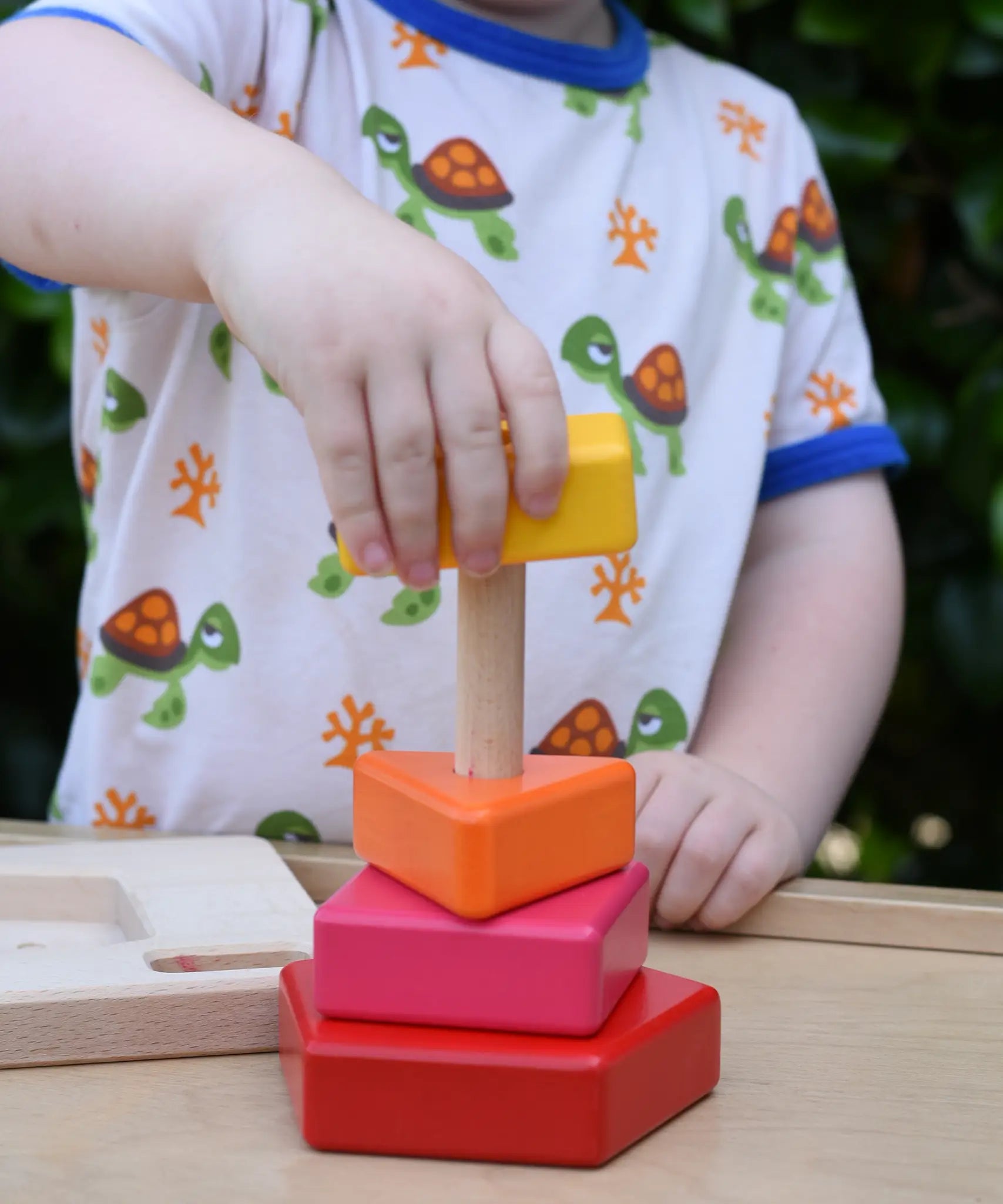 Child playing with the Bajo pyramid sorting board and stacker toy placing a yellow rectangle piece on the stacking pole. 