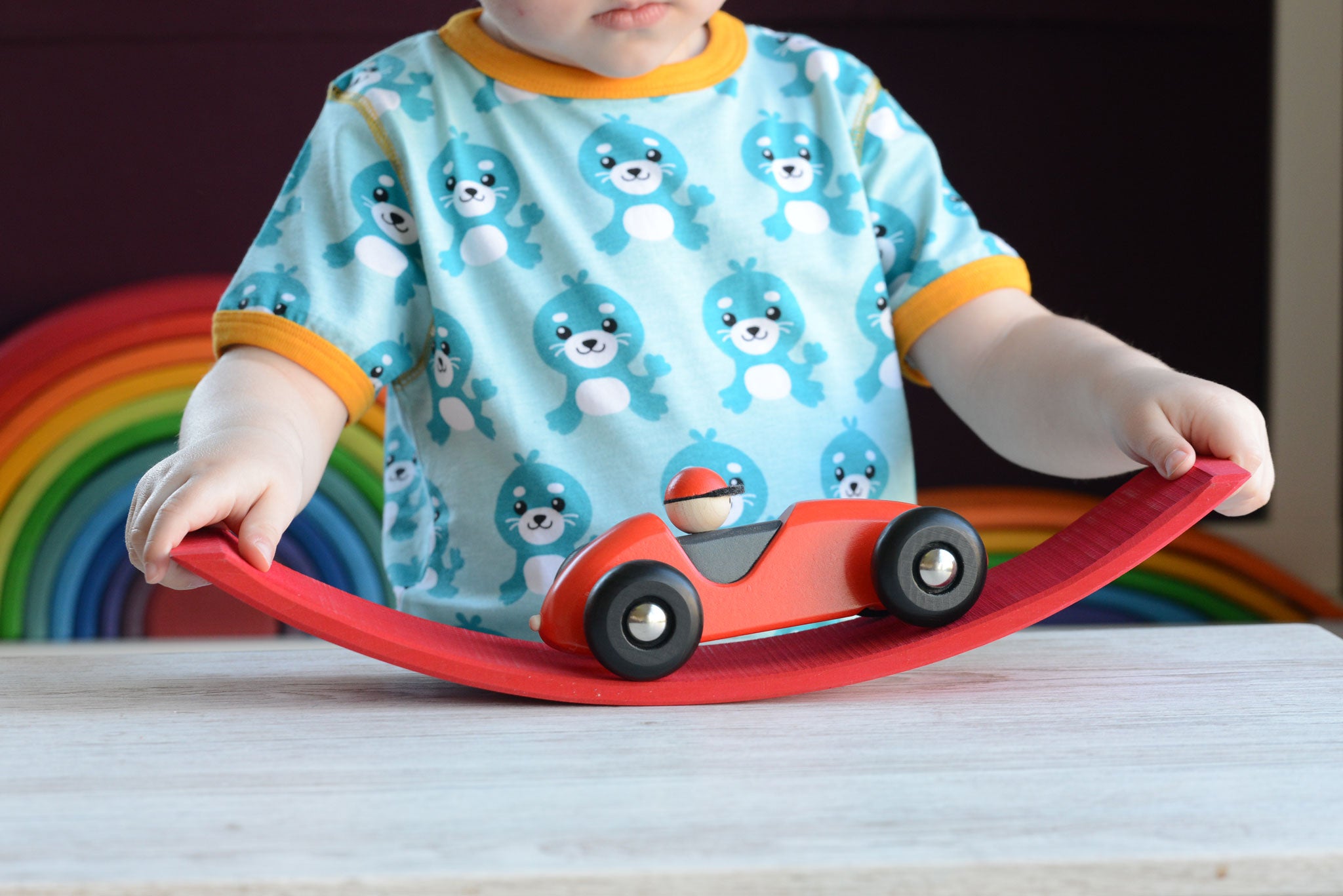 A close up of a child playing with a red Bajo LR car. The child is pushing the car over a red wooden arch. The car is a part of a wide range of Bajo wooden toy vehicles available at Babipur.