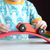 A close up of a child playing with a red Bajo LR car. The child is pushing the car over a red wooden arch. The car is a part of a wide range of Bajo wooden toy vehicles available at Babipur.