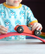 A close up of a child playing with a red Bajo LR car. The child is pushing the car over a red wooden arch. The car is a part of a wide range of Bajo wooden toy vehicles available at Babipur.