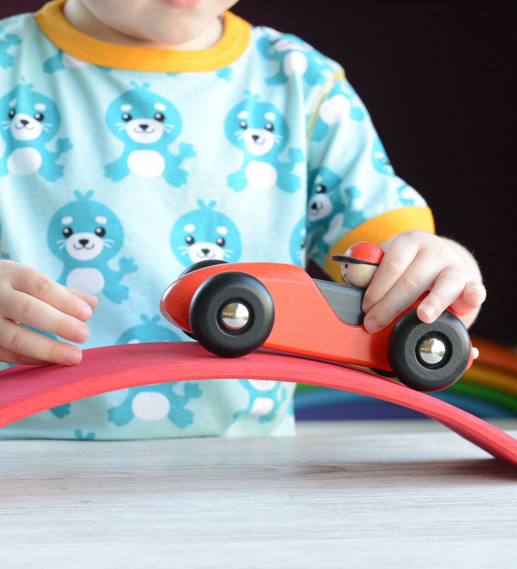 A close up of a child playing with a red Bajo LR car. The child is pushing the car over a red wooden arch. The car is a part of a wide range of Bajo wooden toy vehicles available at Babipur.