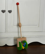 Bajo push along wooden toy rattle leaning against a white cupboard. Green rollers with coloured pieces in the middle 