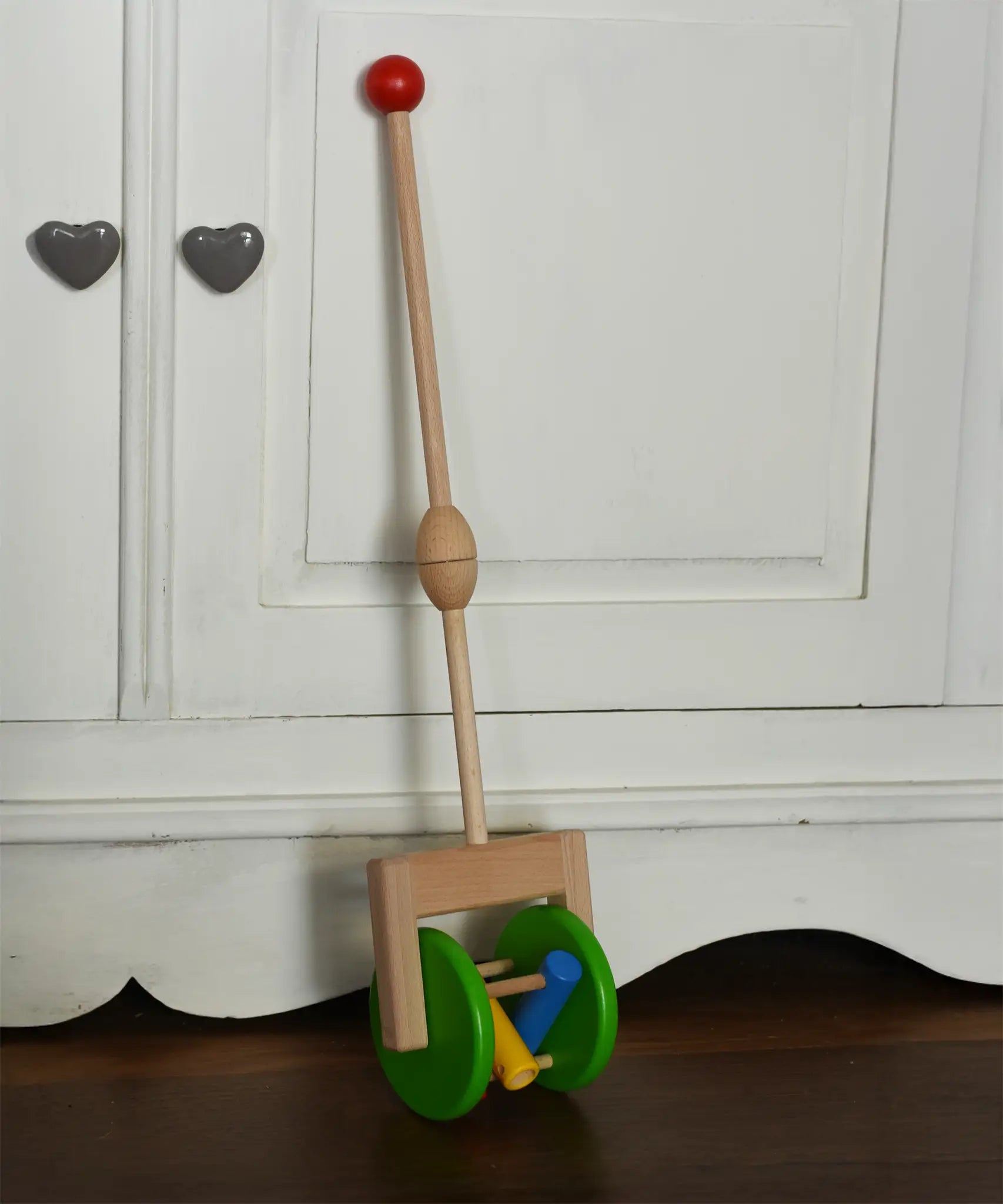 Bajo push along wooden toy rattle leaning against a white cupboard. Green rollers with coloured pieces in the middle 