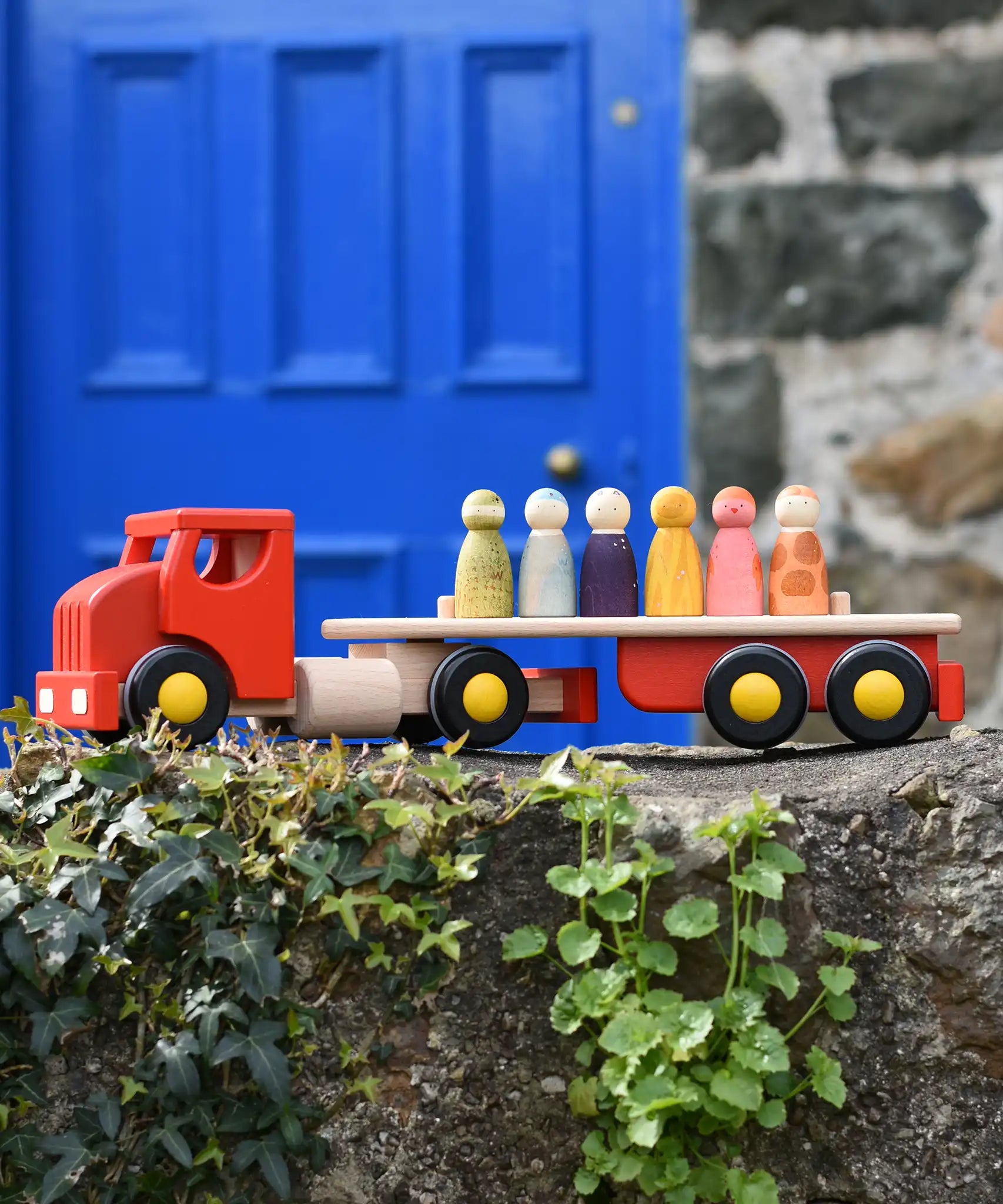 Bajo large red cargo truck and wooden shape sorter toy placed on a stone wall with Grapat wild crew wooden peg dolls placed on the back.