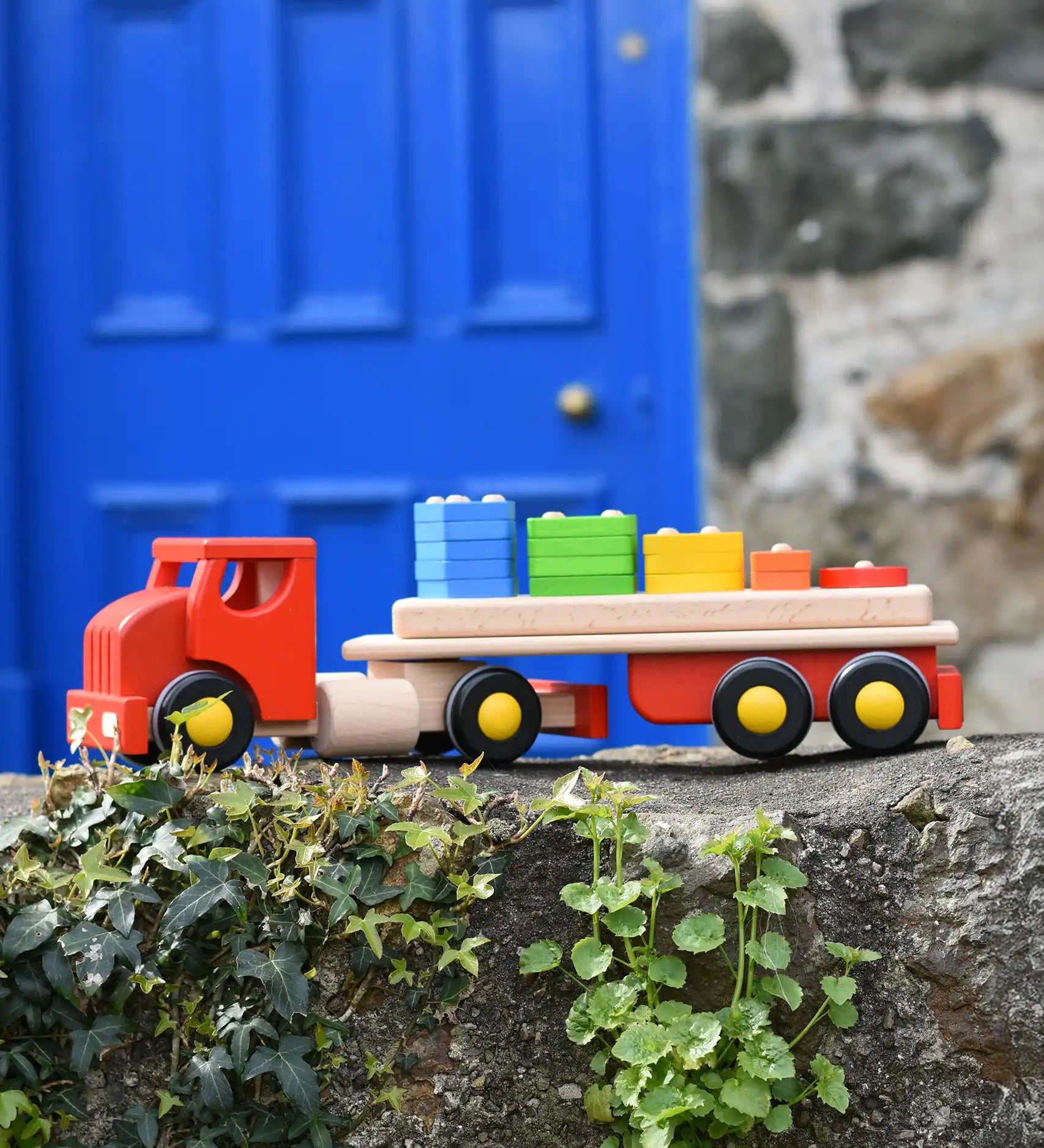 Bajo large red cargo truck and wooden shape sorter toy placed on a stone wall outdoors.