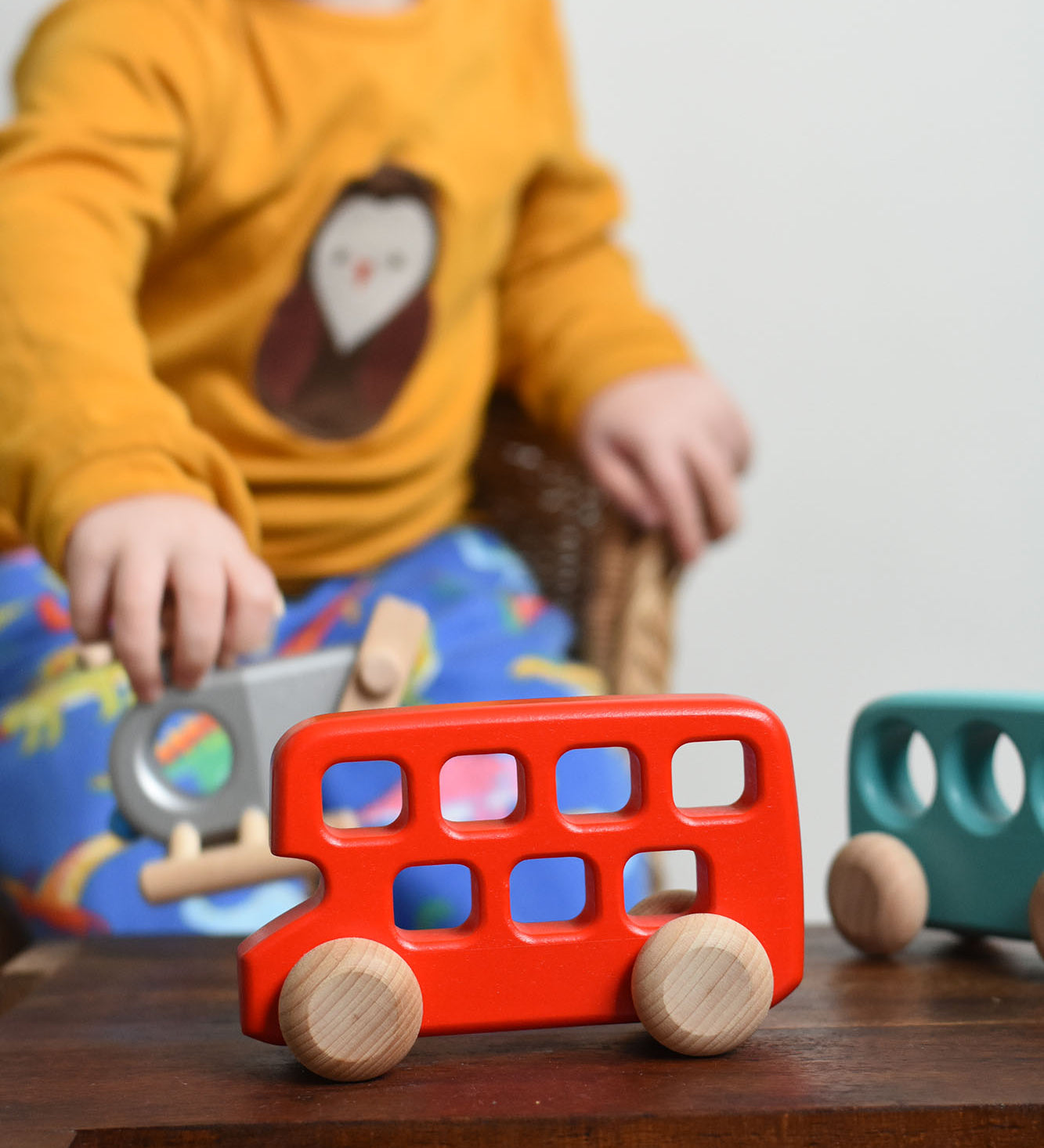 A red coloured Bajo double decker push along wooden toy vehicle placed on a small wooden table. A child can be seen playing with other Bajo wooden toy vehicles. These heirloom quality toys by Bajo are part of a wide range of wooden toys available here at Babipur. 