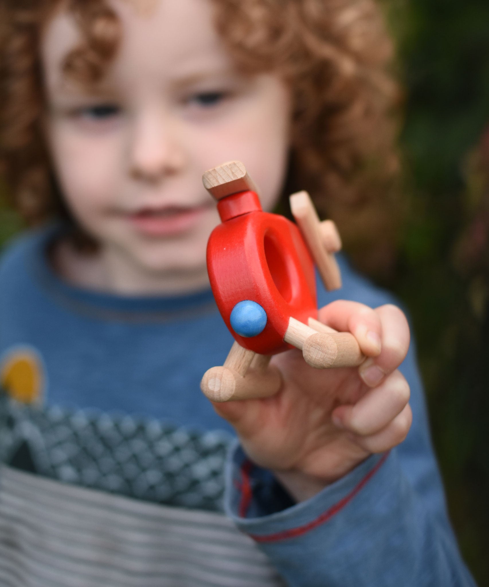 A child holding up a Bajo Red Fire Engine Helicopter wooden toy. The red coloured wooden helicopter has natural propellers and stand and a blue lamp details on the front. These heirloom quality toys by Bajo are part of a wide range of wooden toys available here at Babipur. 