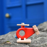 The Bajo Red Fire Engine Helicopter wooden toy placed on a stone wall outdoors. The red coloured wooden helicopter has natural propellers and stand and a blue lamp details on the front. These heirloom quality toys by Bajo are part of a wide range of wooden toys available here at Babipur. 