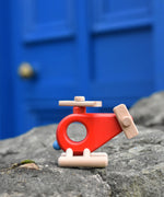 The Bajo Red Fire Engine Helicopter wooden toy placed on a stone wall outdoors. The red coloured wooden helicopter has natural propellers and stand and a blue lamp details on the front. These heirloom quality toys by Bajo are part of a wide range of wooden toys available here at Babipur. 