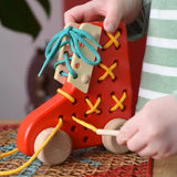 Child pulling the thread on the Bajo red lacing shoe wooden push along toy placed on a rattan stool in the Babipur playroom.
