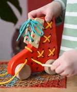 Child pulling the thread on the Bajo red lacing shoe wooden push along toy placed on a rattan stool in the Babipur playroom.
