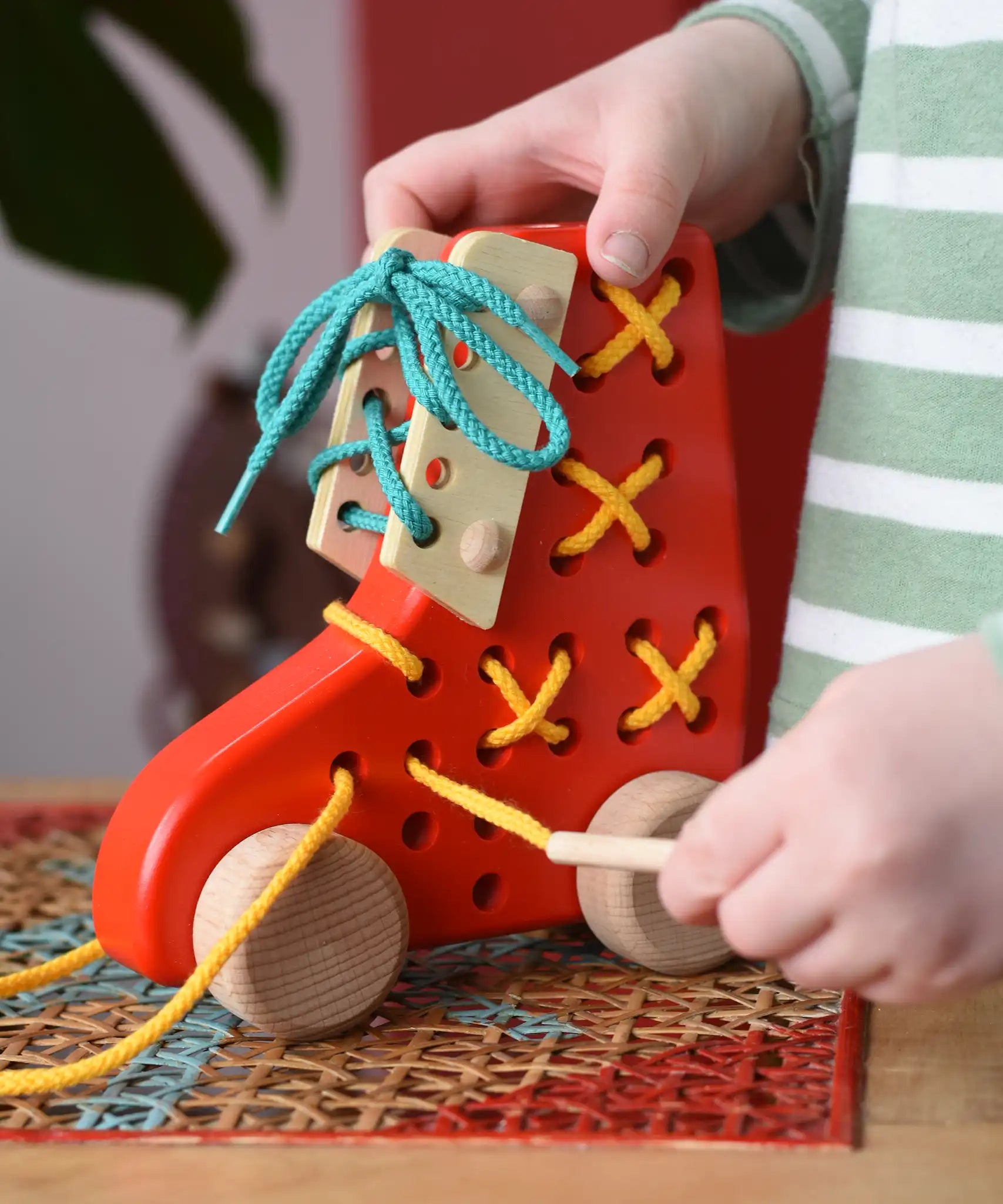 Child pulling the thread on the Bajo red lacing shoe wooden push along toy placed on a rattan stool in the Babipur playroom.