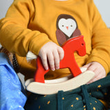A child holding a red coloured Bajo Mini Rocking Horse on their lap. These heirloom quality toys by Bajo are part of a wide range of wooden toys available here at Babipur. 