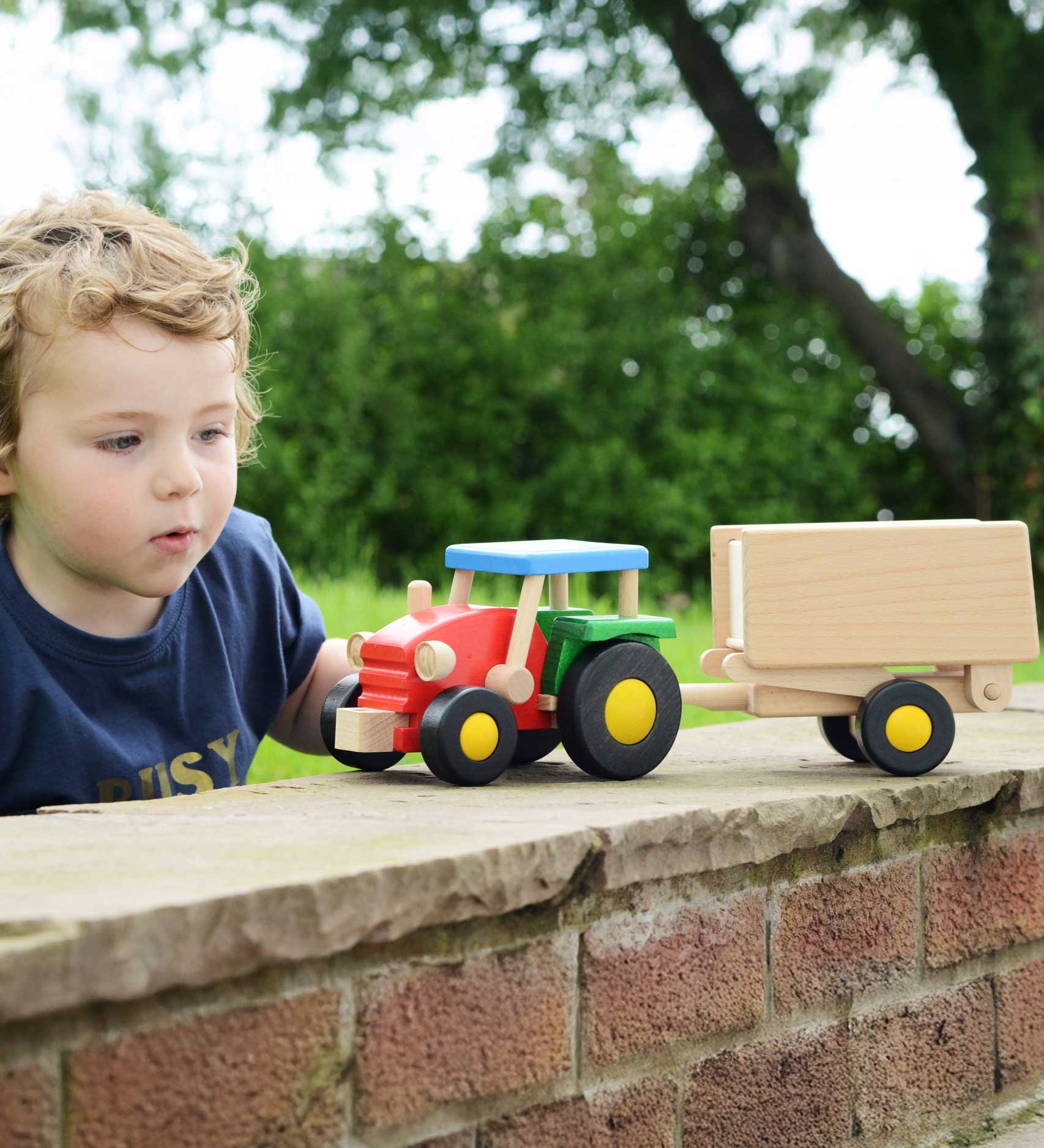 A child playing with the Bajo tractor and trailer outdoors. The tractor has a  red, green and blue painted finish and natural wood trailer. These heirloom quality toys by Bajo are part of a wide range of wooden toys available here at Babipur.