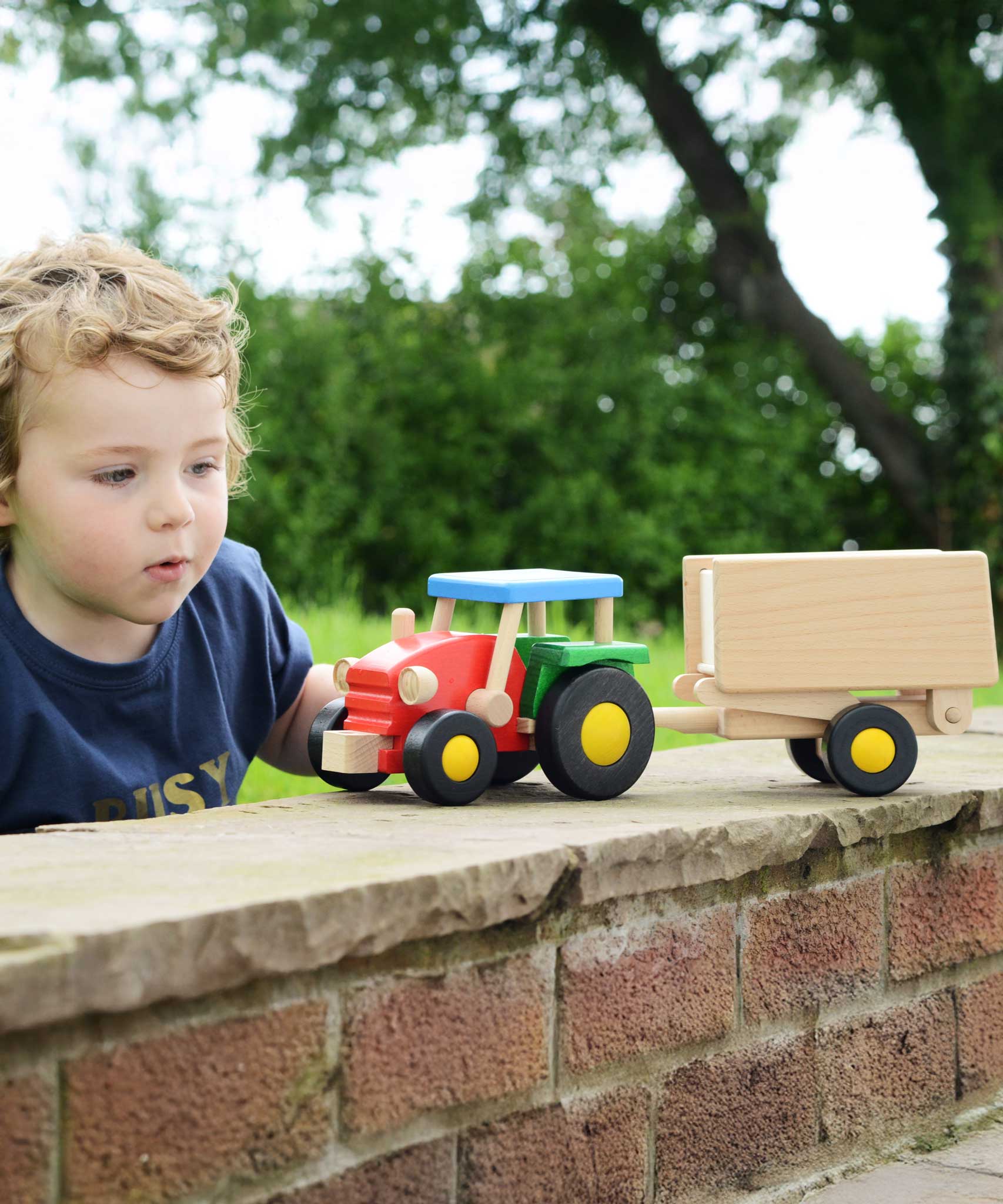 A child playing with the Bajo tractor and trailer outdoors. The tractor has a  red, green and blue painted finish and natural wood trailer. These heirloom quality toys by Bajo are part of a wide range of wooden toys available here at Babipur.