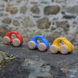 Bajo handmade wooden beetle style car including blue, yellow and red available at Babipur. These have unpainted wheels and can be pushed or pulled along - photo on a stone wall.