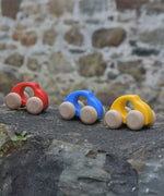 Bajo handmade wooden beetle style car including blue, yellow and red available at Babipur. These have unpainted wheels and can be pushed or pulled along - photo on a stone wall.