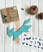 A blue coloured Bajo Tangram puzzle placed on a white wooden surface. The pieces from the Tangram puzzle have been placed in the shape of a fox. The accompanying ideas booklet can be seen open with the cardboard box placed besides it. These heirloom quality toys by Bajo are part of a wide range of wooden toys available here at Babipur. 
