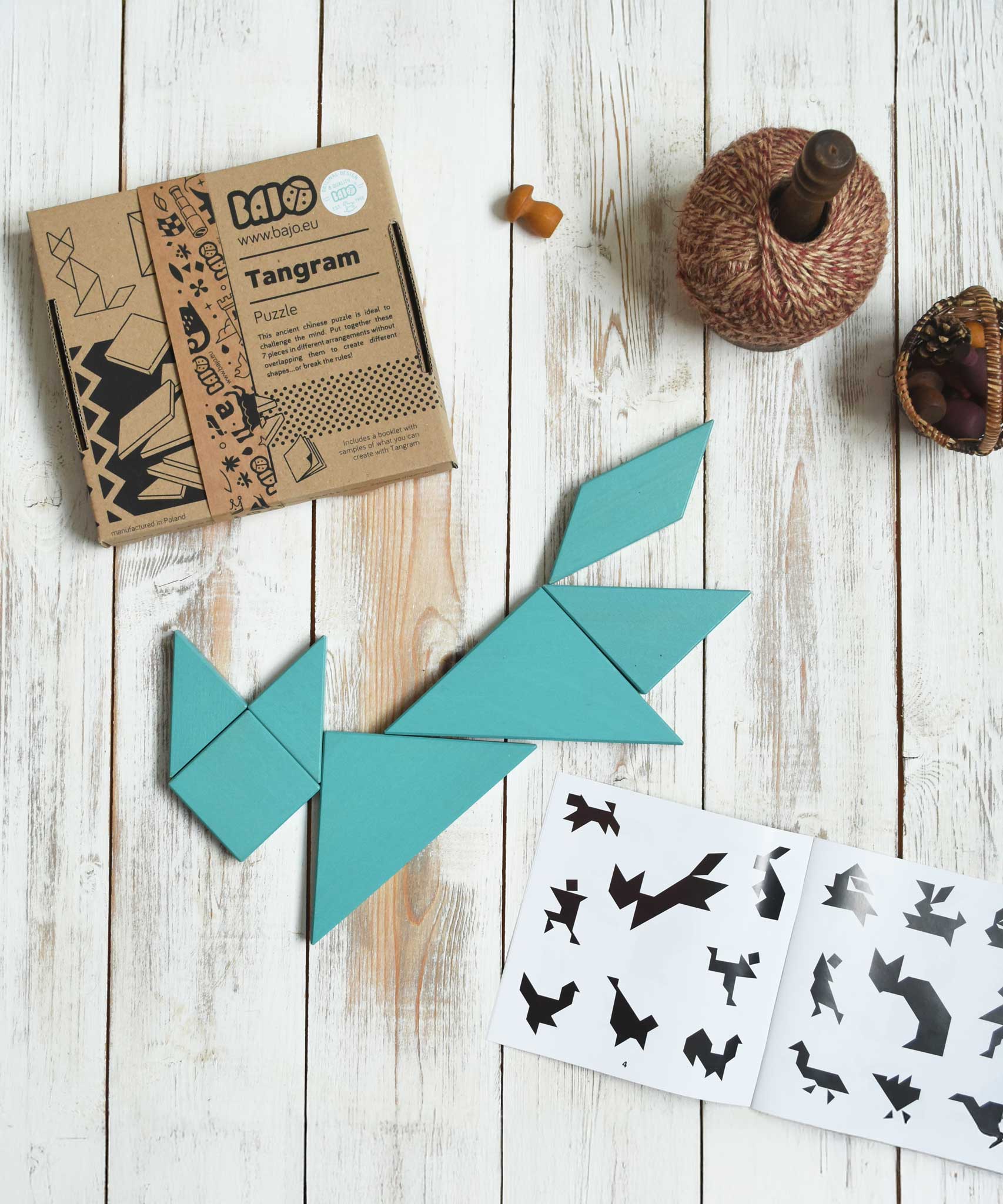 A blue coloured Bajo Tangram puzzle placed on a white wooden surface. The pieces from the Tangram puzzle have been placed in the shape of a fox. The accompanying ideas booklet can be seen open with the cardboard box placed besides it. These heirloom quality toys by Bajo are part of a wide range of wooden toys available here at Babipur. 