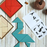 A blue Bajo Tangram puzzle placed on a white wooden surface. The puzzle has been placed in the shape of a bird. The accompanying ideas booklet can be seen open besides it with the red and natural tangram puzzle placed the other side. These heirloom quality toys by Bajo are part of a wide range of wooden toys available here at Babipur. 
