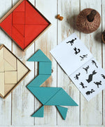 A blue Bajo Tangram puzzle placed on a white wooden surface. The puzzle has been placed in the shape of a bird. The accompanying ideas booklet can be seen open besides it with the red and natural tangram puzzle placed the other side. These heirloom quality toys by Bajo are part of a wide range of wooden toys available here at Babipur. 
